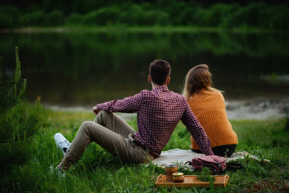 shallow focus photo of man and woman sitting on grass field near body of water during daytime