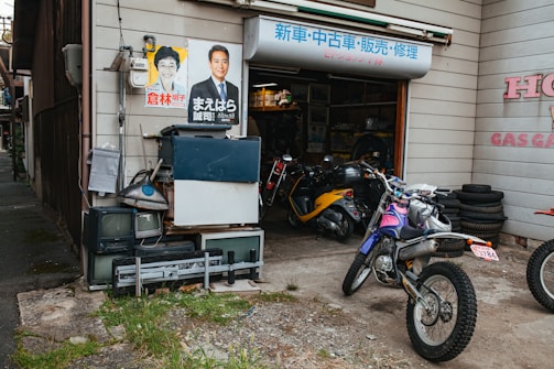 A small motorcycle repair shop with several parked motorbikes is visible. The exterior wall is adorned with posters featuring individuals, and stacked old electronics like televisions and other equipment are present. Piles of tires are neatly organized beside the open garage, where various tools and parts are visible inside.