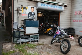 A small motorcycle repair shop with several parked motorbikes is visible. The exterior wall is adorned with posters featuring individuals, and stacked old electronics like televisions and other equipment are present. Piles of tires are neatly organized beside the open garage, where various tools and parts are visible inside.