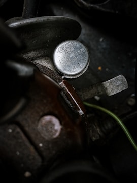 A close-up view of a metallic, round cap on a mechanical device. The cap has inscriptions on it, with the manufacturer name 'Solex' visible. The surrounding area includes parts of machinery with a dark, worn surface, and a greenish cable running alongside.
