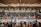 A large indoor arena with a high ceiling, filled with an audience seated on bleachers. In the center, two fighters are engaged in a martial arts competition on blue and red mats. The scene is captured through a mesh fence, adding a textured foreground.