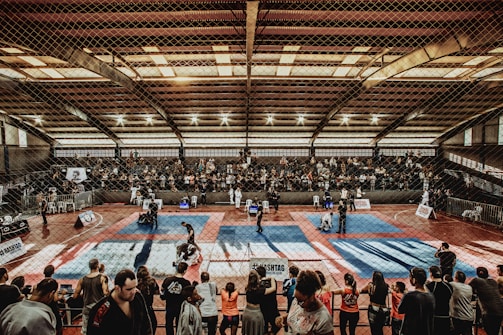 A large indoor arena with a high ceiling, filled with an audience seated on bleachers. In the center, two fighters are engaged in a martial arts competition on blue and red mats. The scene is captured through a mesh fence, adding a textured foreground.