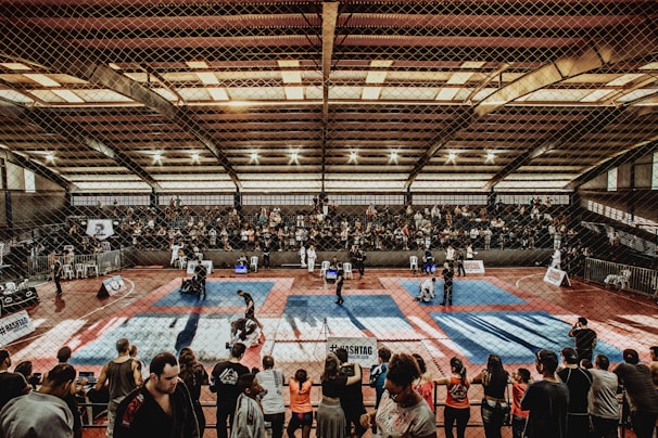 A large indoor arena with a high ceiling, filled with an audience seated on bleachers. In the center, two fighters are engaged in a martial arts competition on blue and red mats. The scene is captured through a mesh fence, adding a textured foreground.