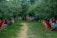A serene outdoor setting with a path leading through a grove of trees, surrounded by numerous red deck chairs occupied by people. Some individuals are engaged in conversation or relaxing, creating a communal and laid-back atmosphere.