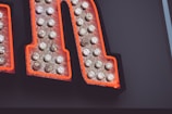 A close-up view of a section of a large letter, outlined in neon orange with numerous exposed light bulbs on its surface, likely part of a vintage or retro sign.