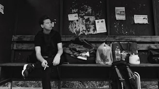 A young person is sitting on a weathered wooden bench inside a shelter, holding a cigarette. Various bags, a blanket, and a kettle are placed beside them. The wall behind is peeling and features several torn posters.
