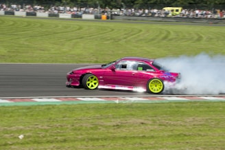 A bright pink sports car with neon green wheels drifts on a racetrack, emitting thick white smoke from the rear tires. A crowd of spectators is visible in the background, observing the event. The racetrack is bordered by neatly trimmed grass.