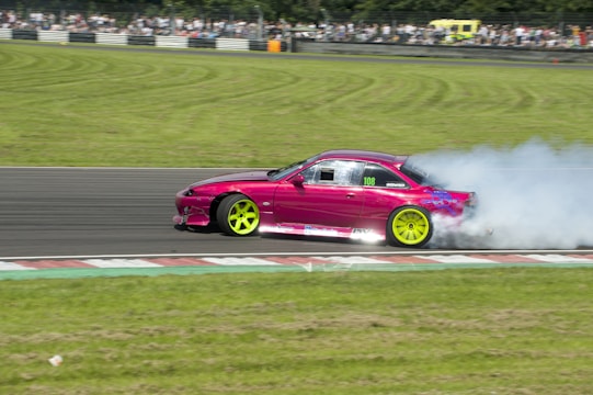 A bright pink sports car with neon green wheels drifts on a racetrack, emitting thick white smoke from the rear tires. A crowd of spectators is visible in the background, observing the event. The racetrack is bordered by neatly trimmed grass.