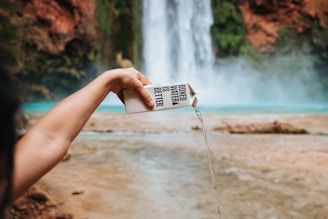 A hand is pouring water from a carton labeled 'Boxed Water is Better' in front of a blurred waterfall with a pool at the base. The background consists of a natural landscape with water and rugged terrain.