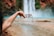 A hand is pouring water from a carton labeled 'Boxed Water is Better' in front of a blurred waterfall with a pool at the base. The background consists of a natural landscape with water and rugged terrain.
