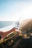 A person holds a carton labeled 'Boxed Water Is Better' with water splashing out, set against a scenic background of a coastal cliff and ocean under clear blue skies. The sunlight casts a warm glow, creating a serene and invigorating atmosphere.