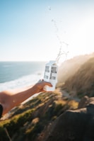 A person holds a carton labeled 'Boxed Water Is Better' with water splashing out, set against a scenic background of a coastal cliff and ocean under clear blue skies. The sunlight casts a warm glow, creating a serene and invigorating atmosphere.
