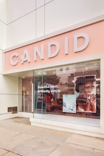 A store facade with a large sign reading 'CANDID' features a modern, bright design. The window display includes promotional text that states 'Beautiful teeth. Brilliantly simple.' There is a large image of a person holding a dental product. The interior seems uncluttered with chic and minimalist furniture visible through the glass.