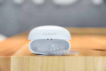A transparent dental aligner is partially visible inside a white case with the brand name 'Candid' on it. The case rests on a wooden surface with a blurred background.