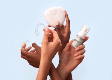 Several hands hold dental care items including a clear dental aligner, a whitening product in a small bottle, and a storage case labeled 'Candid'. The background is a solid light blue, providing a clean and minimalistic appearance.