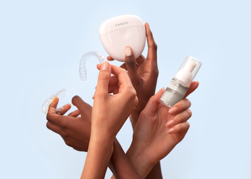 Several hands hold dental care items including a clear dental aligner, a whitening product in a small bottle, and a storage case labeled 'Candid'. The background is a solid light blue, providing a clean and minimalistic appearance.