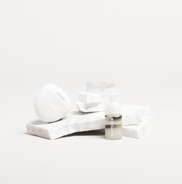 A white, sleek container with the brand name 'Candid', a small bottle labeled 'Whitener', and a clear dental aligner rest on marble slabs. The composition is minimalist with an emphasis on dental care products.