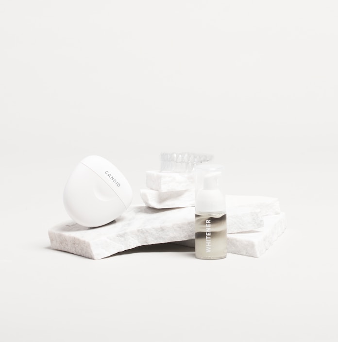 A white, sleek container with the brand name 'Candid', a small bottle labeled 'Whitener', and a clear dental aligner rest on marble slabs. The composition is minimalist with an emphasis on dental care products.