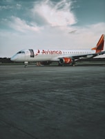 A commercial airplane from Avianca is stationed on a vast tarmac under a partly cloudy sky. The aircraft has a white body with red accents, including the tail and engines. The foreground shows an expansive concrete surface, while the background features a line of trees under a calm sky.