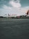 A commercial airplane from Avianca is stationed on a vast tarmac under a partly cloudy sky. The aircraft has a white body with red accents, including the tail and engines. The foreground shows an expansive concrete surface, while the background features a line of trees under a calm sky.