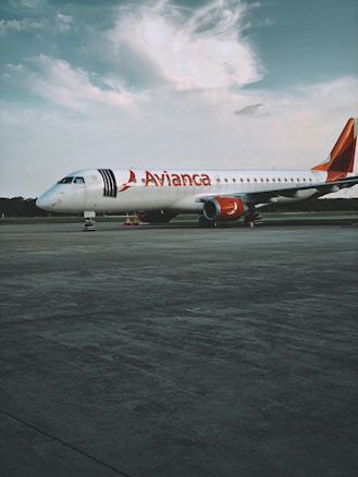A commercial airplane from Avianca is stationed on a vast tarmac under a partly cloudy sky. The aircraft has a white body with red accents, including the tail and engines. The foreground shows an expansive concrete surface, while the background features a line of trees under a calm sky.