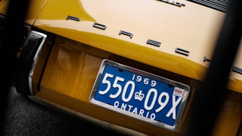 A close-up view of the rear end of a yellow Porsche car, displaying a blue Ontario license plate from 1969. The car's logo is prominently displayed above the license plate.