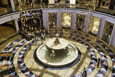An elegantly designed indoor space featuring a central water fountain surrounded by a circular, geometric-patterned floor. The scene is illuminated by numerous hanging cylindrical lights, creating a warm and inviting atmosphere. In the background, there are retail store entrances with ornate displays. People are casually walking around the area, adding a sense of hustle and bustle.