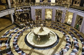 An elegantly designed indoor space featuring a central water fountain surrounded by a circular, geometric-patterned floor. The scene is illuminated by numerous hanging cylindrical lights, creating a warm and inviting atmosphere. In the background, there are retail store entrances with ornate displays. People are casually walking around the area, adding a sense of hustle and bustle.