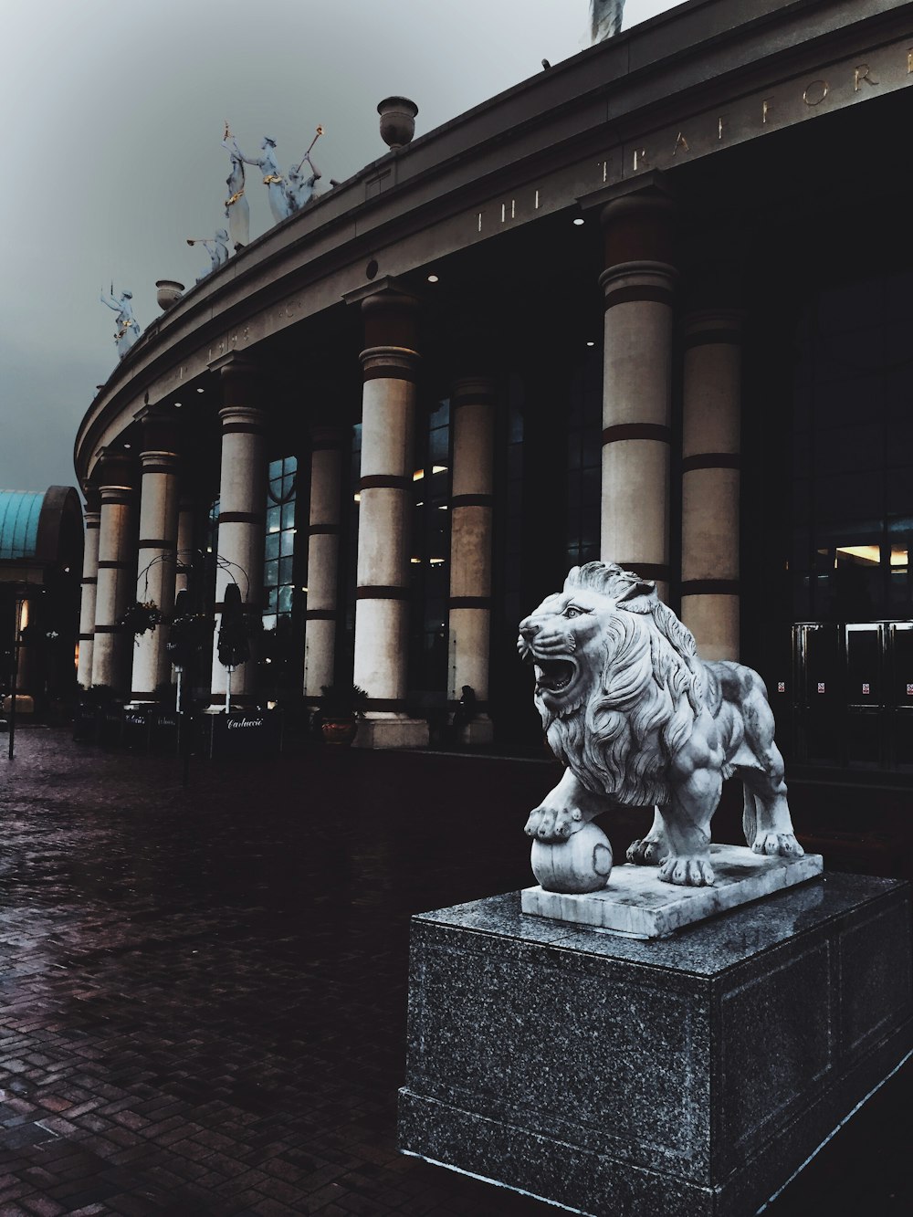 Statue de lion adulte sur une base en béton devant un bâtiment avec des piliers extérieurs