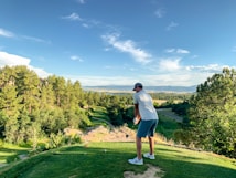 A person is preparing to swing a golf club on a golf course, surrounded by lush green trees under a clear blue sky. The landscape extends into the distance with a view of mountains and a winding road.