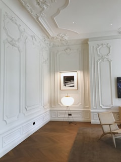 A sophisticated and elegant room features detailed white wall moldings and a decorative ceiling. A modern lamp with a spherical shade stands on a wooden floor, accompanied by a woven chair on a beige rug. A framed black and white photograph is mounted on the wall.