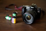 A Canon camera with a large lens is placed on a wooden surface. In front of the camera, there are three film rolls in different colors, indicating different types and brands. The film rolls have labels displaying '100 T-Max' and 'Fujifilm', suggesting traditional photographic processes.