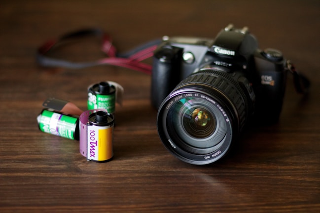 A Canon camera with a large lens is placed on a wooden surface. In front of the camera, there are three film rolls in different colors, indicating different types and brands. The film rolls have labels displaying '100 T-Max' and 'Fujifilm', suggesting traditional photographic processes.