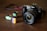 A Canon camera with a large lens is placed on a wooden surface. In front of the camera, there are three film rolls in different colors, indicating different types and brands. The film rolls have labels displaying '100 T-Max' and 'Fujifilm', suggesting traditional photographic processes.
