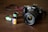 A Canon camera with a large lens is placed on a wooden surface. In front of the camera, there are three film rolls in different colors, indicating different types and brands. The film rolls have labels displaying '100 T-Max' and 'Fujifilm', suggesting traditional photographic processes.