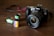 A Canon camera with a large lens is placed on a wooden surface. In front of the camera, there are three film rolls in different colors, indicating different types and brands. The film rolls have labels displaying '100 T-Max' and 'Fujifilm', suggesting traditional photographic processes.