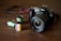A Canon camera with a large lens is placed on a wooden surface. In front of the camera, there are three film rolls in different colors, indicating different types and brands. The film rolls have labels displaying '100 T-Max' and 'Fujifilm', suggesting traditional photographic processes.