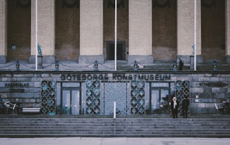 A grand entrance to a museum with a name displayed in large letters. The facade features stone steps leading up to double glass doors. Decorative elements include metalwork and sculptures. Two people stand near the entrance amidst the stone architecture.