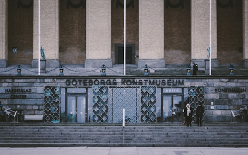 A grand entrance to a museum with a name displayed in large letters. The facade features stone steps leading up to double glass doors. Decorative elements include metalwork and sculptures. Two people stand near the entrance amidst the stone architecture.