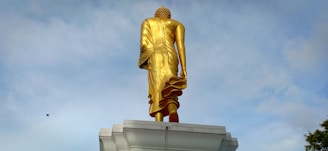 A large golden statue of Buddha is viewed from the back, positioned on a white platform against a cloudy blue sky. The statue is towering and detailed, capturing the folds of the garments and the textured hairstyle.