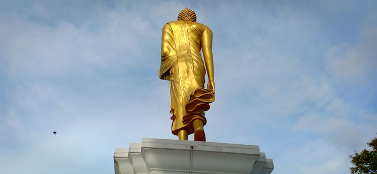 A large golden statue of Buddha is viewed from the back, positioned on a white platform against a cloudy blue sky. The statue is towering and detailed, capturing the folds of the garments and the textured hairstyle.