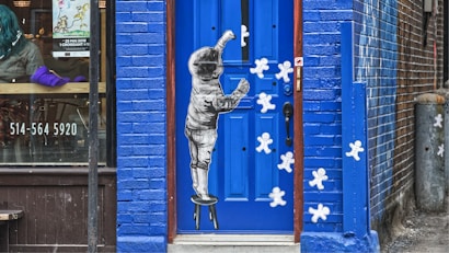 A blue door with an artistic depiction of a child on a stool reaching upwards, surrounded by white gingerbread man shapes on the door. The door is set within a vibrant blue-painted brick wall. To the left, part of a cafe window is visible with a person inside, along with text showing a phone number and an event poster.