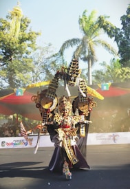 An individual is dressed in an elaborate, traditional costume adorned with intricate details, gold and red accents, including a detailed headpiece. The background displays a festival atmosphere with colorful banners and trees under a clear sky, suggesting an outdoor event.