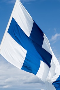 A flag with a blue Nordic cross on a white background waves against a bright, partly cloudy sky.