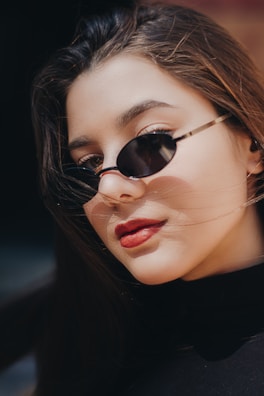 A close-up of a person wearing dark sunglasses and red lipstick. The lighting emphasizes smooth skin and flowing dark hair, suggesting a fashionable and confident posture.