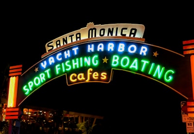 A brightly lit, colorful neon sign with the words 'Santa Monica Yacht Harbor Sport Fishing Boating Cafes' against a dark background.