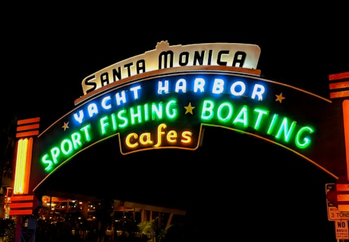 A brightly lit, colorful neon sign with the words 'Santa Monica Yacht Harbor Sport Fishing Boating Cafes' against a dark background.
