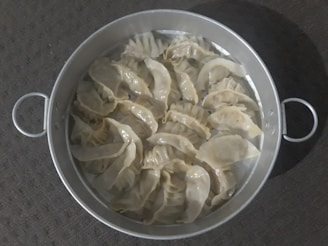 A round metal steamer filled with several steamed dumplings arranged neatly. The dumplings have a glossy, slightly translucent appearance, indicating they are freshly cooked. The steamer has two handles, and the background features a textured, dark surface.