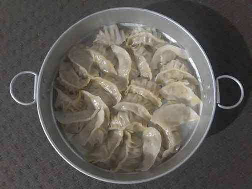 A round metal steamer filled with several steamed dumplings arranged neatly. The dumplings have a glossy, slightly translucent appearance, indicating they are freshly cooked. The steamer has two handles, and the background features a textured, dark surface.