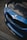 A close-up view of the front part of a blue BMW car, showcasing the iconic BMW emblem and grille. The hood appears to have a carbon fiber finish, and there are reflections visible on the surface. The logo of KZ Auto Group is visible on the bottom part of the bumper.